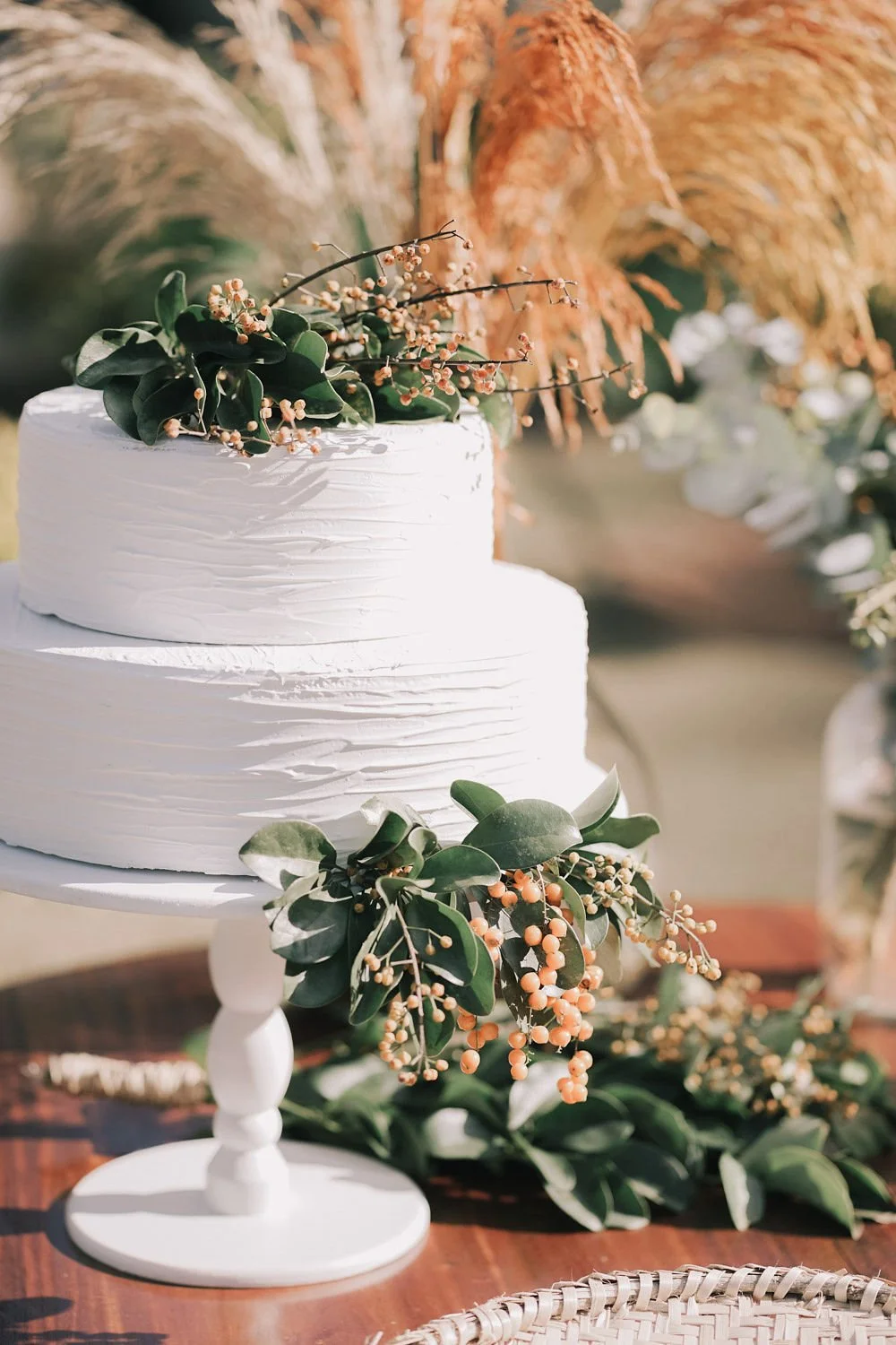 Bolo de casamento branco decorado com folhagens verdes e pequenos frutos, sobre um suporte branco, em ambiente de cerimônia ao ar livre.
