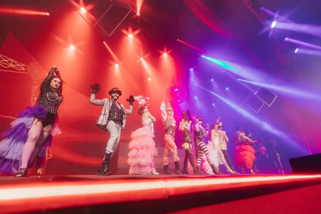 Vários artistas de circo em uma apresentação no palco usando roupas coloridas, com iluminação de luzes coloridas e efeitos de luz.