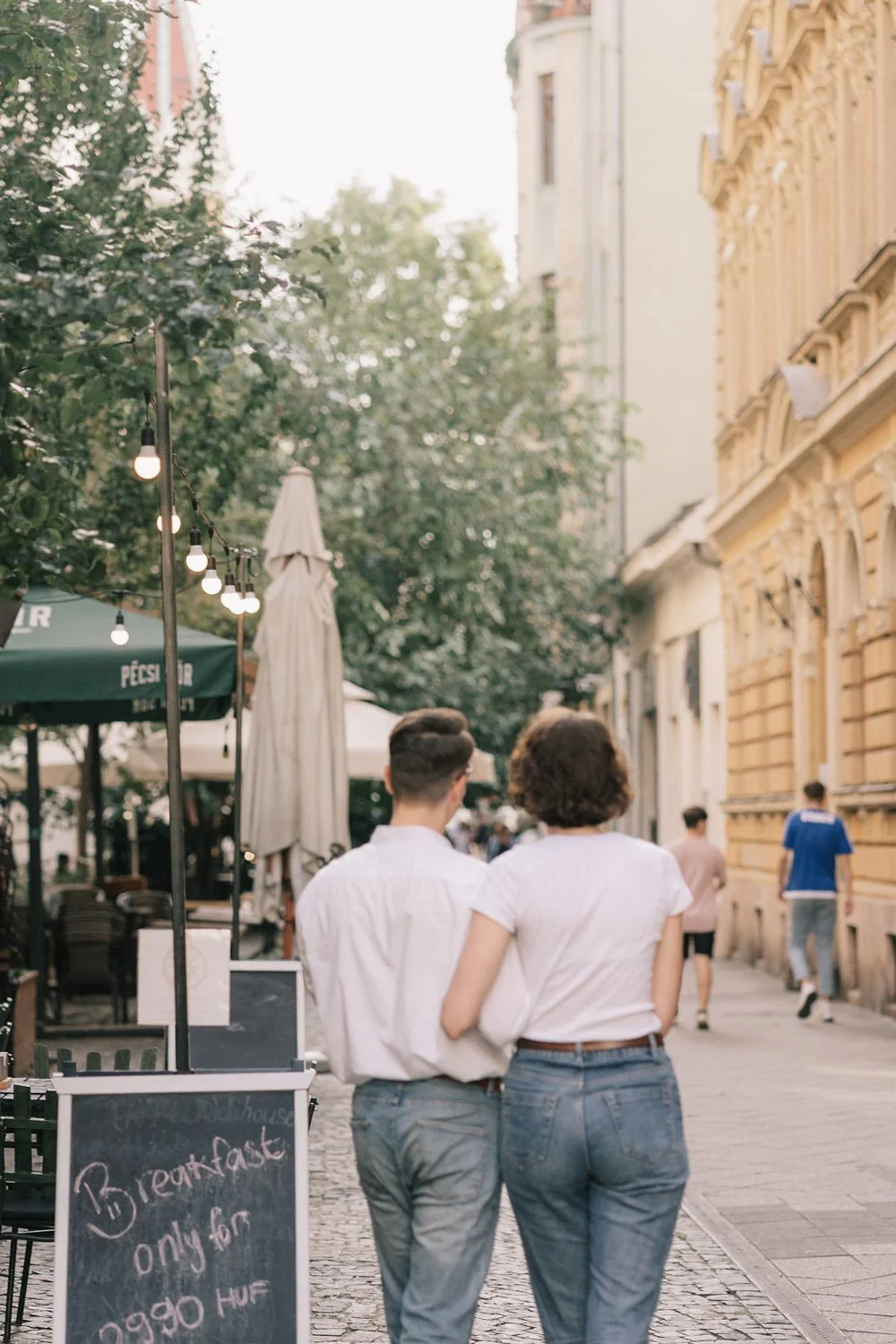Casal caminhando pelo calçadão de uma rua com edifícios antigos, árvores e iluminação de rua com lâmpadas expostas.