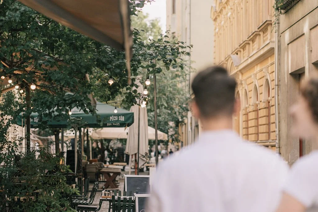 Pessoa com cabelo curto e outra com cabelo loiro, vista de costas, em uma rua ao ar livre com mesas de um café, árvores e edifícios antigos ao fundo.