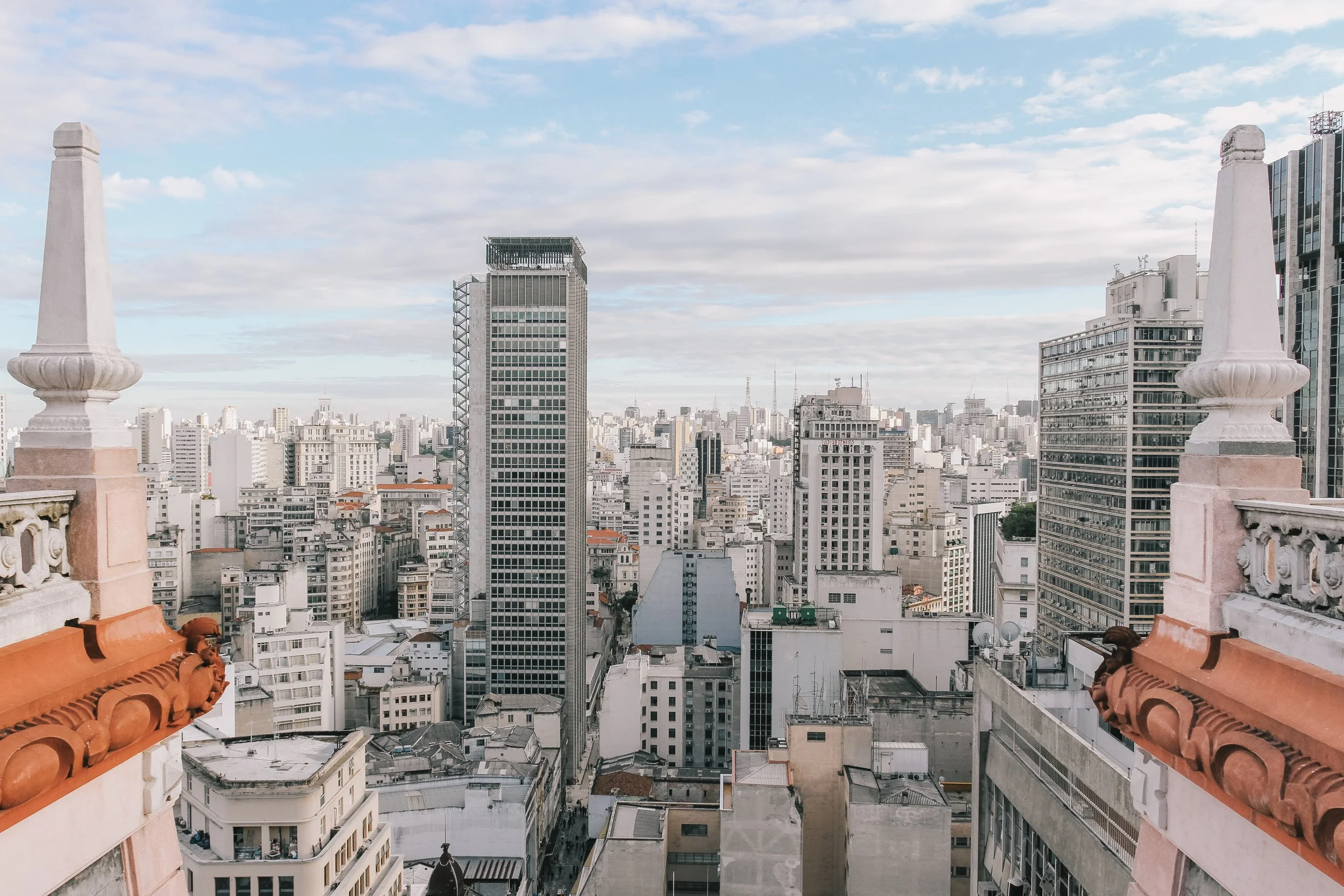 Vista panorâmica de uma cidade com prédios altos, céu com algumas nuvens e elementos arquitetônicos antigos na varanda