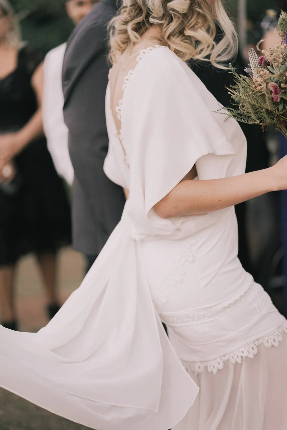 Imagem de uma noiva vestida de branco, segurando um buquê de flores, em um casamento ao ar livre.