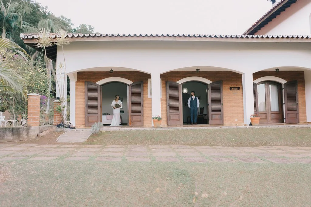 Casarão com três portas de madeira, uma mulher vestida de branco e um homem de terno azul escuro na varanda.