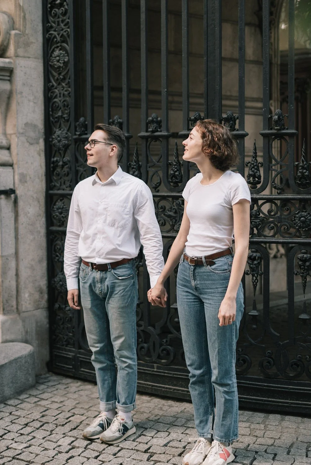 Homem e mulher segurando as mãos em frente a uma cerca preta de ferro, sorrindo e se olhando, na rua de paralelepípedos.