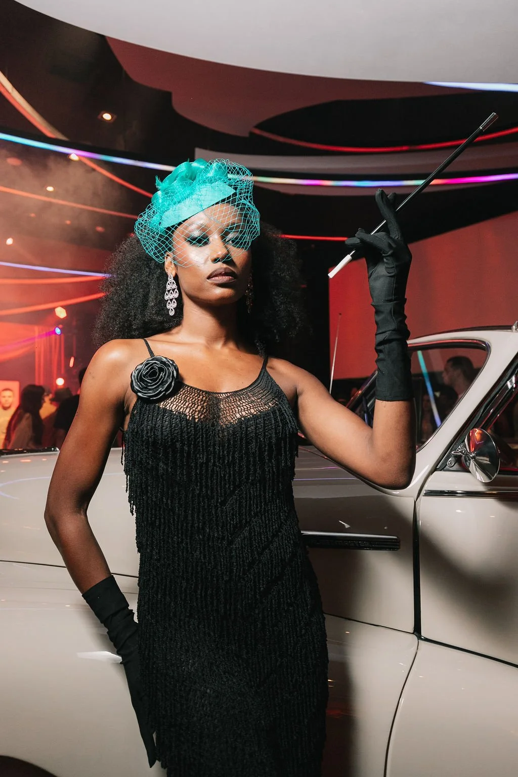 Mulher elegante com vestido preto, luvas longas pretas, acessório de cabeça azul, penteado com cabelo cacheado, segurando um cachimbo, ao lado de um carro branco, em ambiente com luzes coloridas.