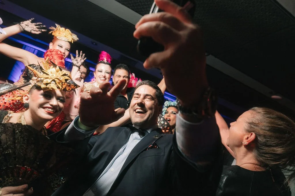 Grupo de pessoas sorrindo e posando para uma selfie em uma festa, algumas vestindo fantasias coloridas com acessórios elaborados.