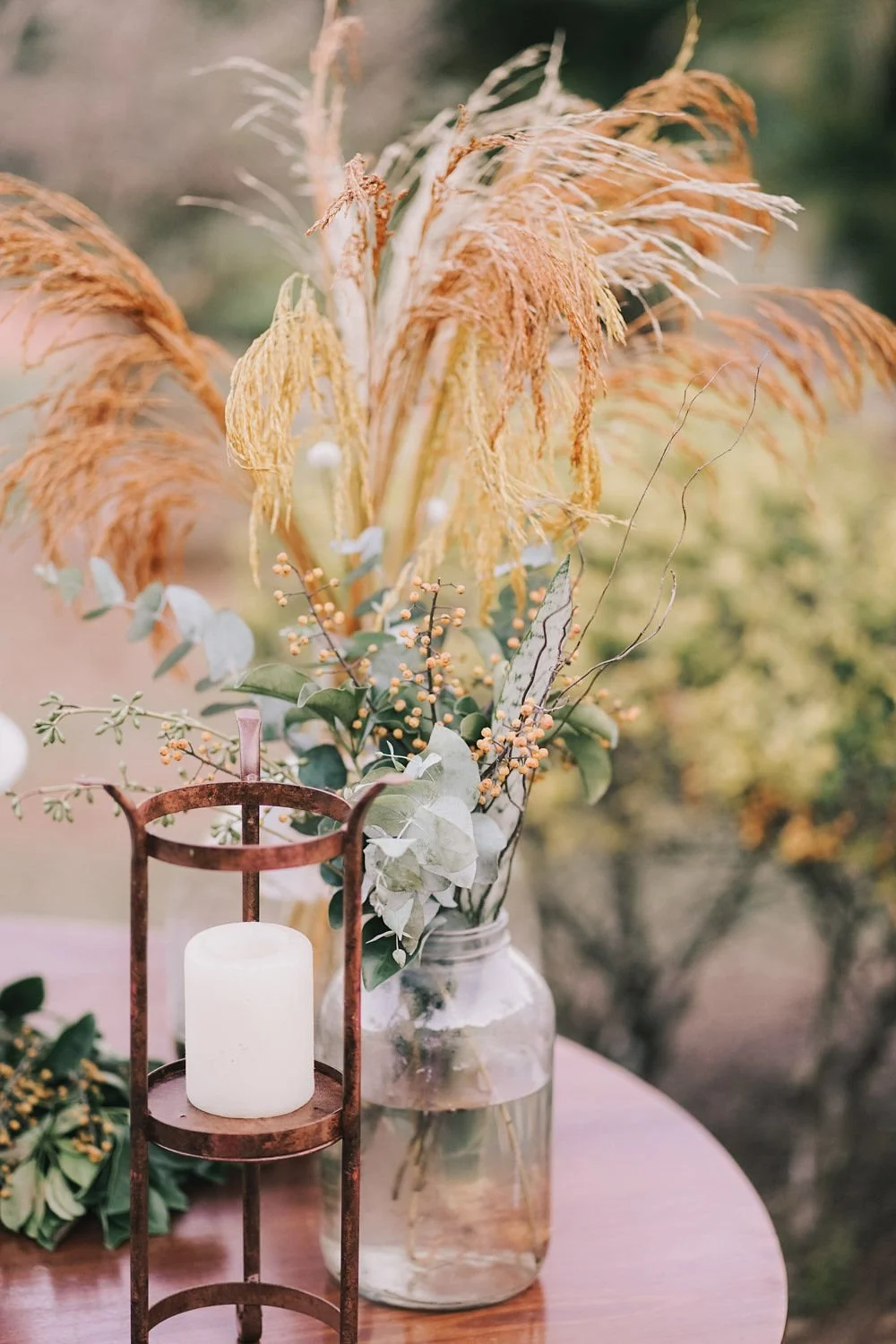 Arranjo de flores com folhas verdes, galhos com pequenas bolinhas, e plantas secas com folhas amarelas e alvas em um recipiente de vidro. Ao lado, uma vela branca dentro de uma estrutura de ferro no topo de uma mesa de madeira.