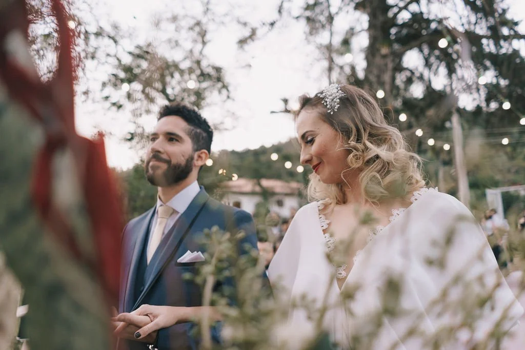 Casal em casamento ao ar livre, noivo e noiva sorrindo, árvores ao fundo, luzes decorativas, atmosfera romântica.
