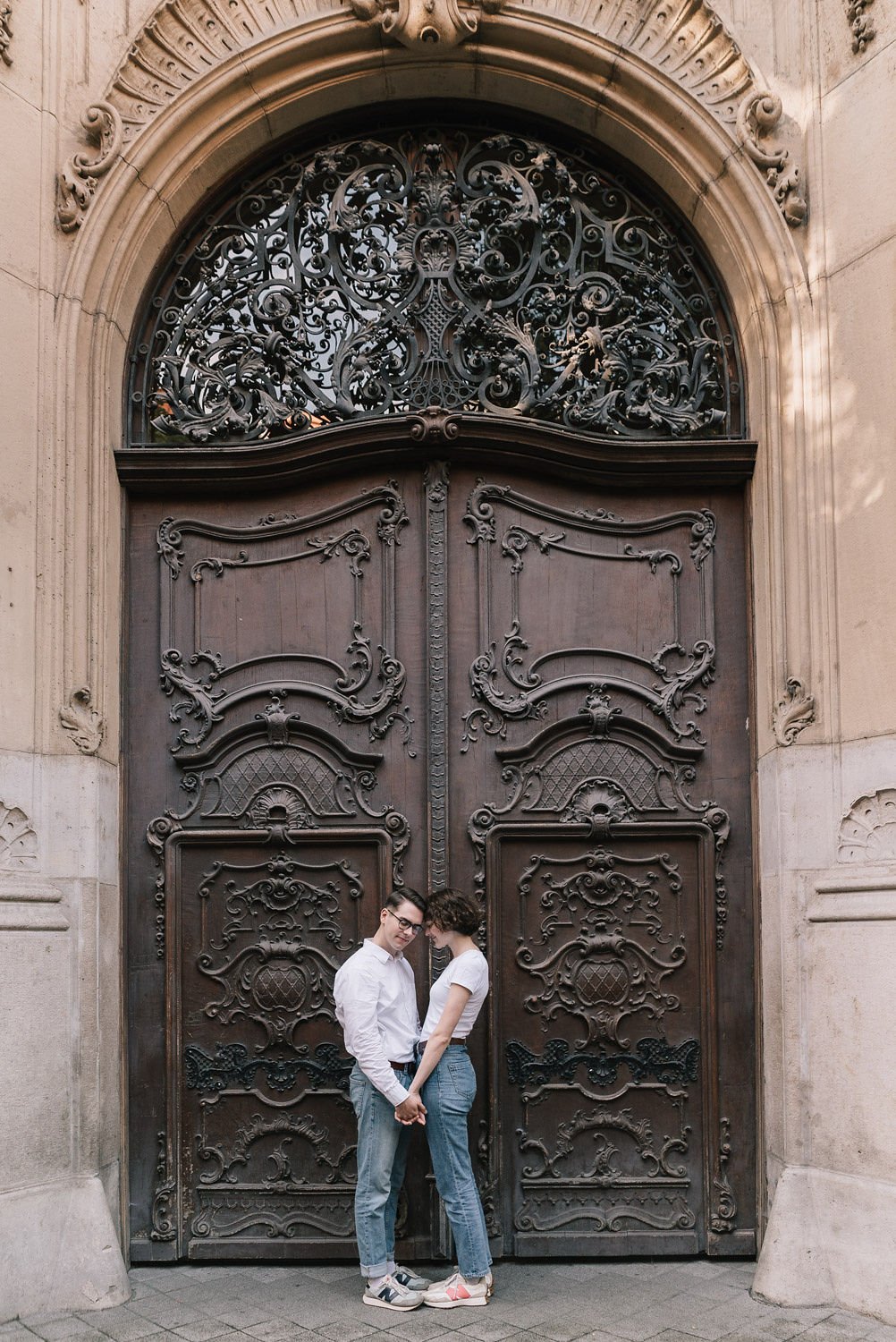 Casal segurando as mãos na frente de uma grande porta de metal ornamentada e decorada com detalhes elegantes, situada em uma estrutura de pedra.