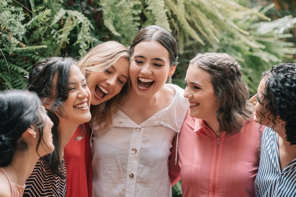 Grupo de mulheres sorrindo e se abraçando ao ar livre, com vegetação verde ao fundo.
