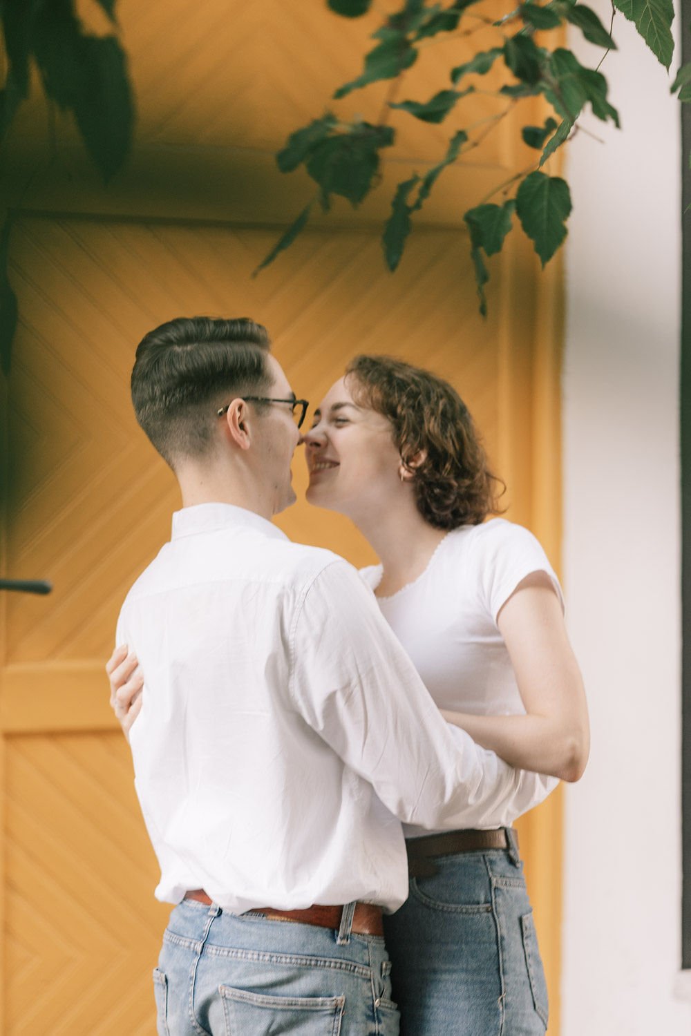 Um casal sorridente se abraçando, quase se beijando, em um ambiente com paredes de madeira e plantas penduradas.
