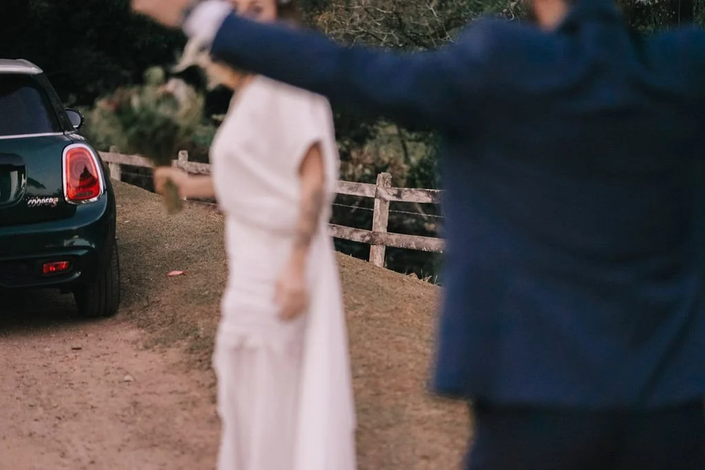 Pessoa com roupa branca segurando uma rosa ao lado de um carro azul escuro, enquanto outra pessoa com roupa azul estende o braço.