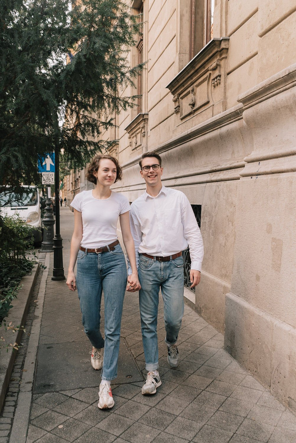Casal jovem caminhando de mãos dadas na calçada de uma rua urbana, sorrindo e usando roupas casuais brancas e jeans, com uma árvore verde e edifícios históricos ao fundo.