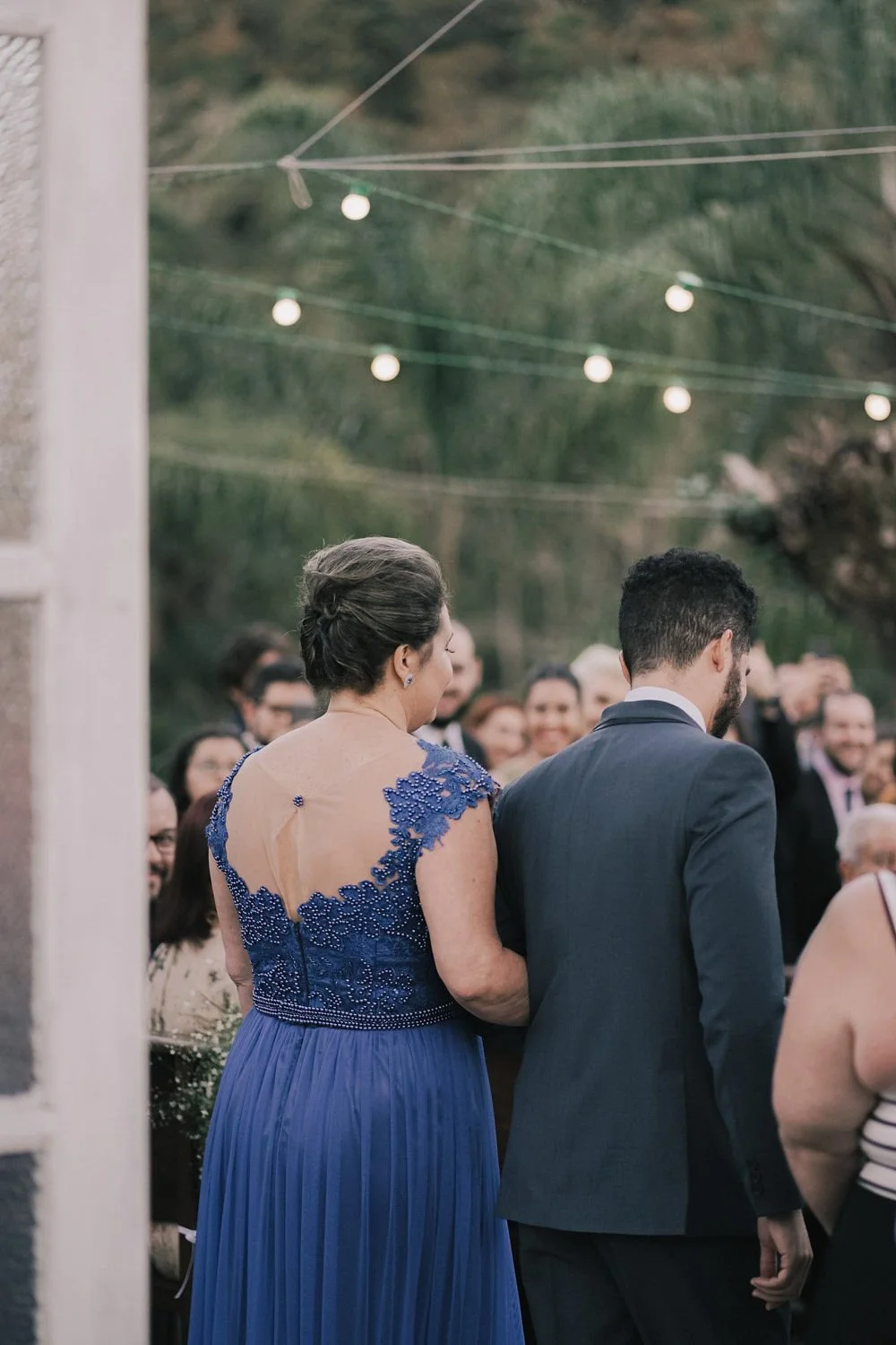 Casamento ao ar livre com convidados. Mulher de vestido azul com detalhes de renda na parte de cima e homem de terno preto ao lado dela, entre convidados sorridentes.