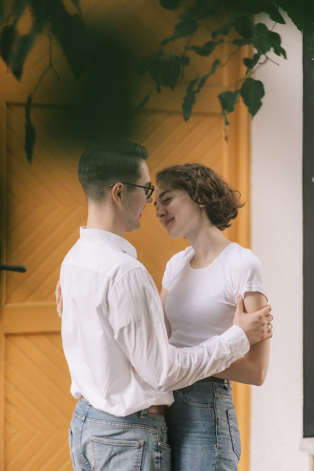 Um casal jovem sorrindo e se abraçando, de frente um para o outro, em um ambiente com paredes de madeira e folhas de plantas visíveis na parte superior da imagem.