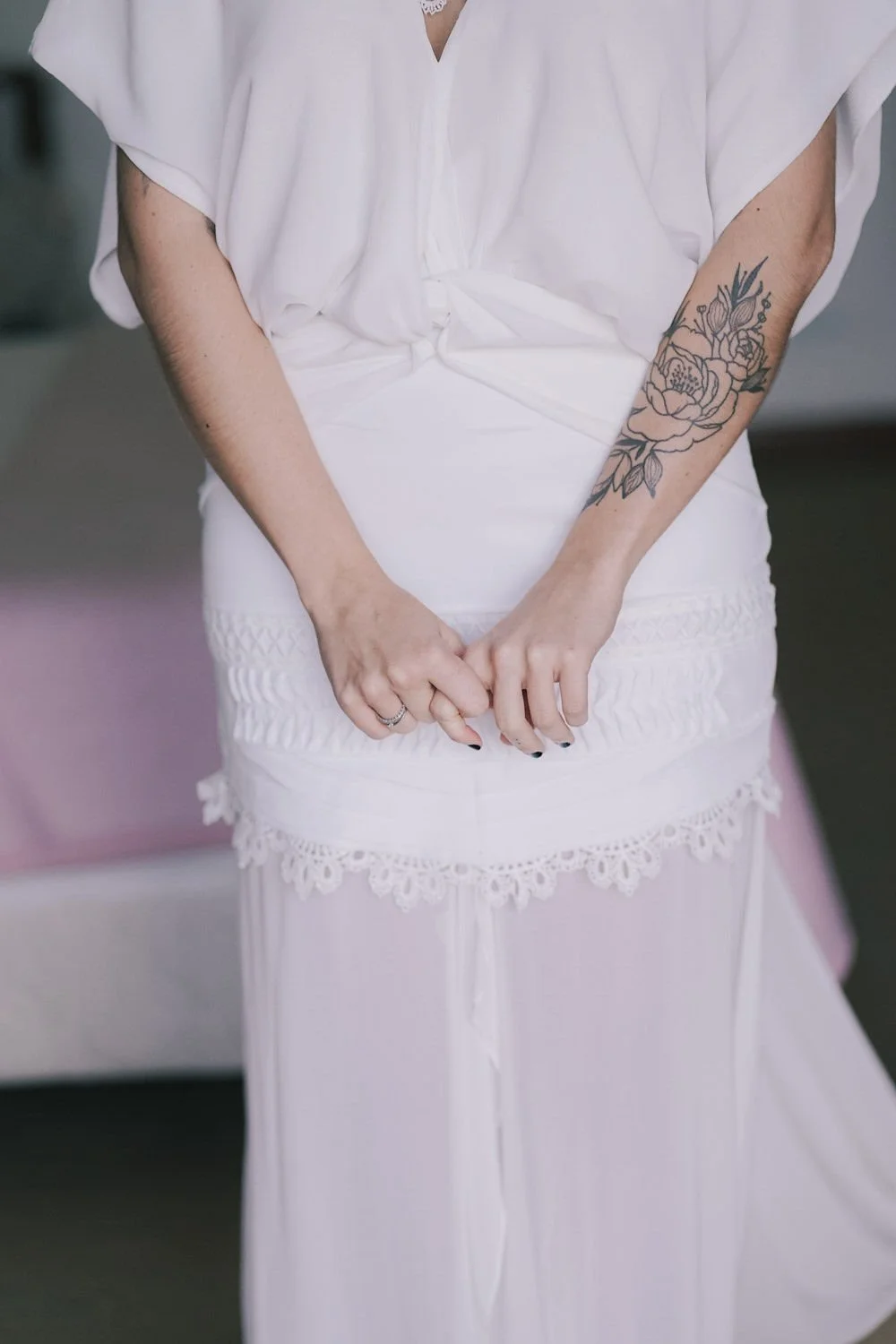 Pessoa usando vestido branco de renda com tatuagem de flor no braço direito, as mãos entrelaçadas na frente do vestido.