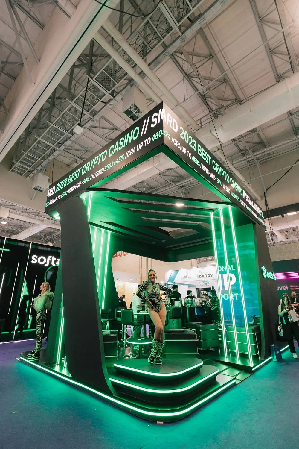 Estande de feira com iluminação verde, promocional de cripto moedas, pessoas dentro, uma mulher posando para foto usando roupa e botas de cano alto em frente ao balcão.