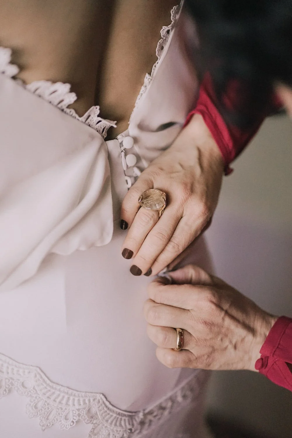Mãos de duas pessoas, uma com anel grande na mão, segurando a roupa de casamento com detalhes em renda.