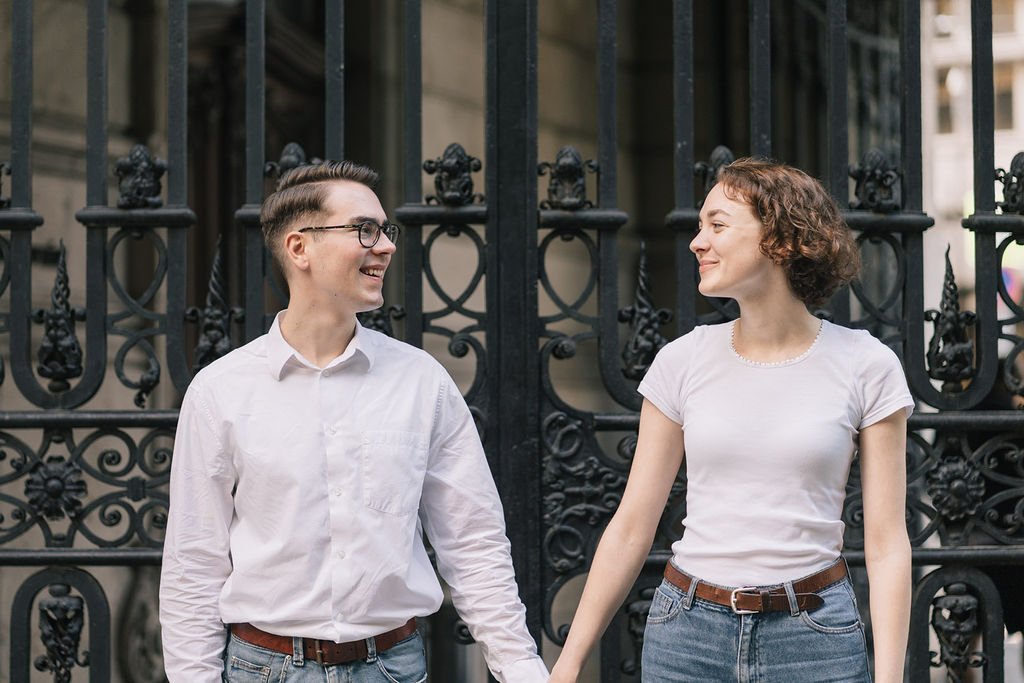 Um casal jovem sorrindo, de mãos dadas, na rua, na frente de um portão de ferro decorado.