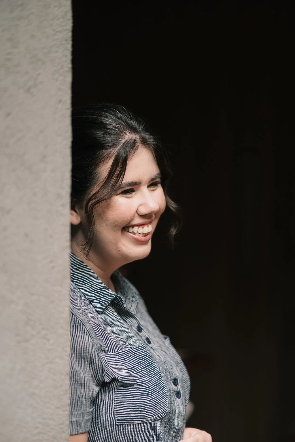 Primeira mulher sorrindo, olhando para o lado, em um ambiente com fundo preto, usando uma blusa de botões com padrão listrado.