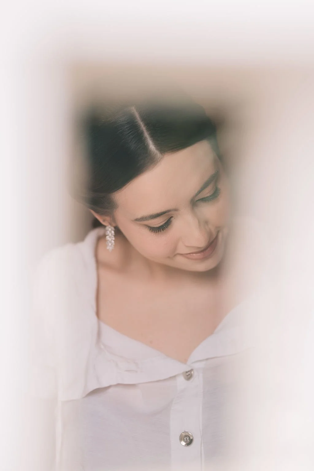 Mulher com cabelo escuro, penteado clássico, usando brincos de pérola, vestindo blusa branca com botões, sorrindo com os olhos fechados.