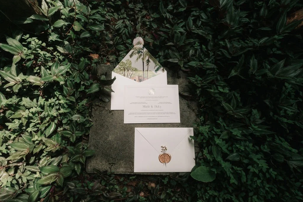 Convite de casamento sobre uma pedra, cercado por folhagens verdes, com envelope e detalhes decorativos