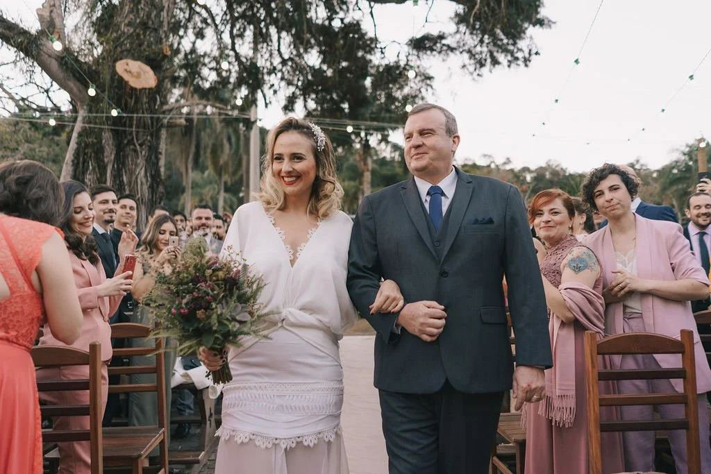 Casal de noivos caminhando na cerimônia de casamento ao ar livre, com convidados sorrindo ao redor e árvores ao fundo.