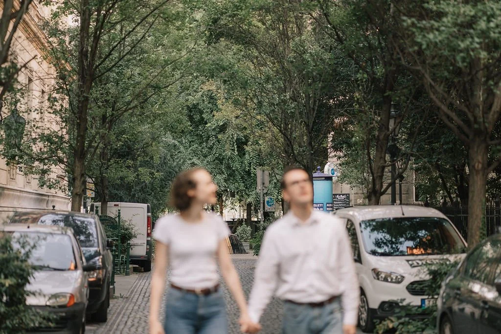 Casal jovem caminhando de mãos dadas na rua, com árvores e carros estacionados ao redor