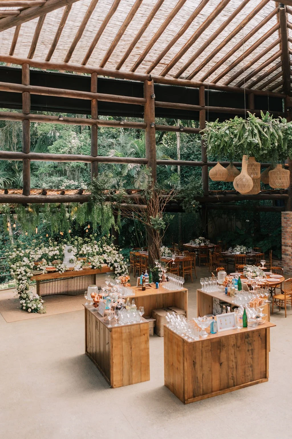 Decoração de evento ao ar livre com mesas de madeira, flores brancas, muitas plantas verdes e cactos pendurados, em um espaço com estrutura de madeira no teto e uma parede de vidro com vista para a natureza.