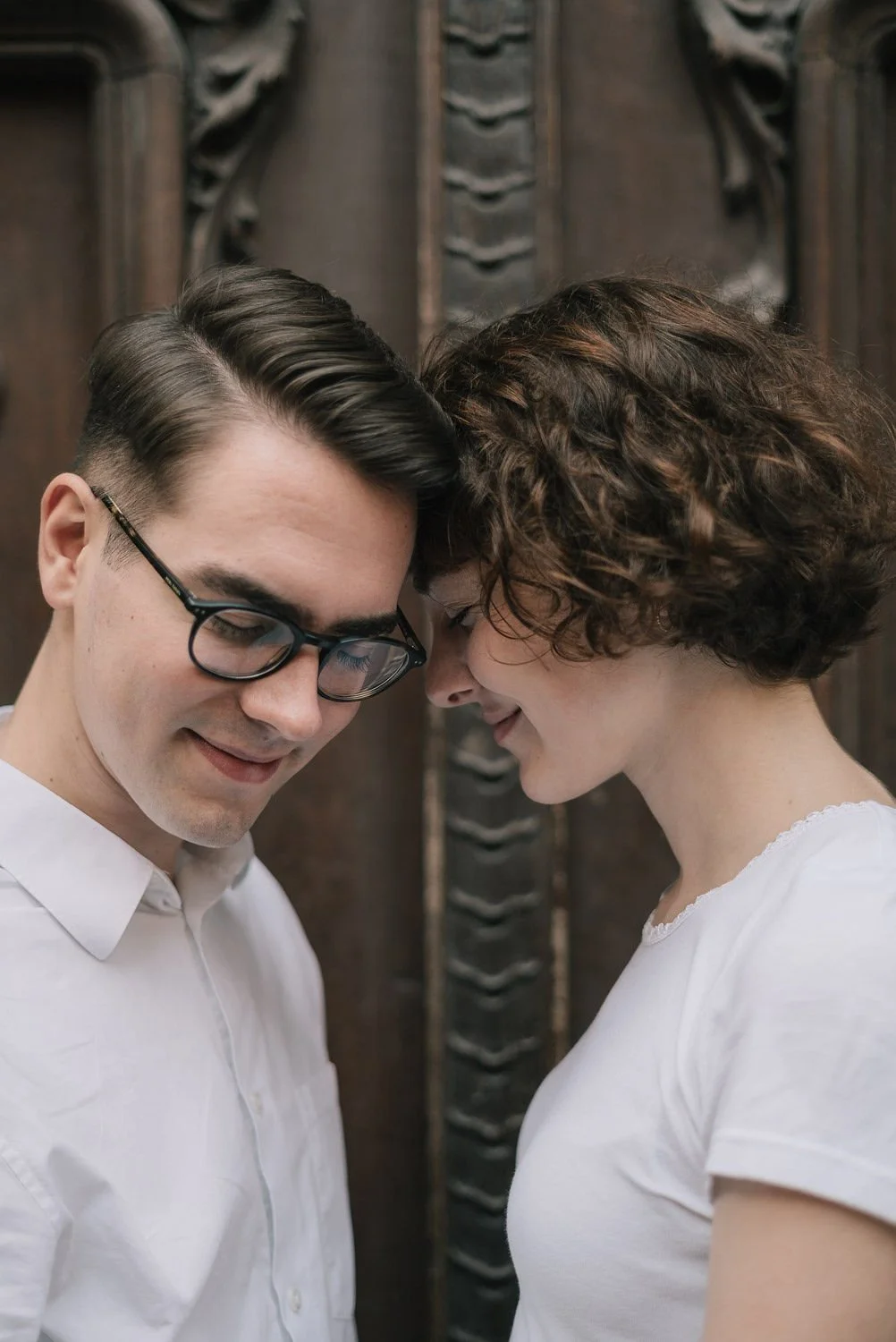 Homem com óculos e mulher com cabelo cacheado, ambos de cabeça baixa e com testa encostada, sorrindo e tocando testa numa parede de madeira.