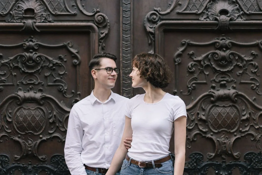 Um casal jovem de mãos dadas, sorrindo e olhando um ao outro, em frente a uma porta de madeira decorada com detalhes artísticos