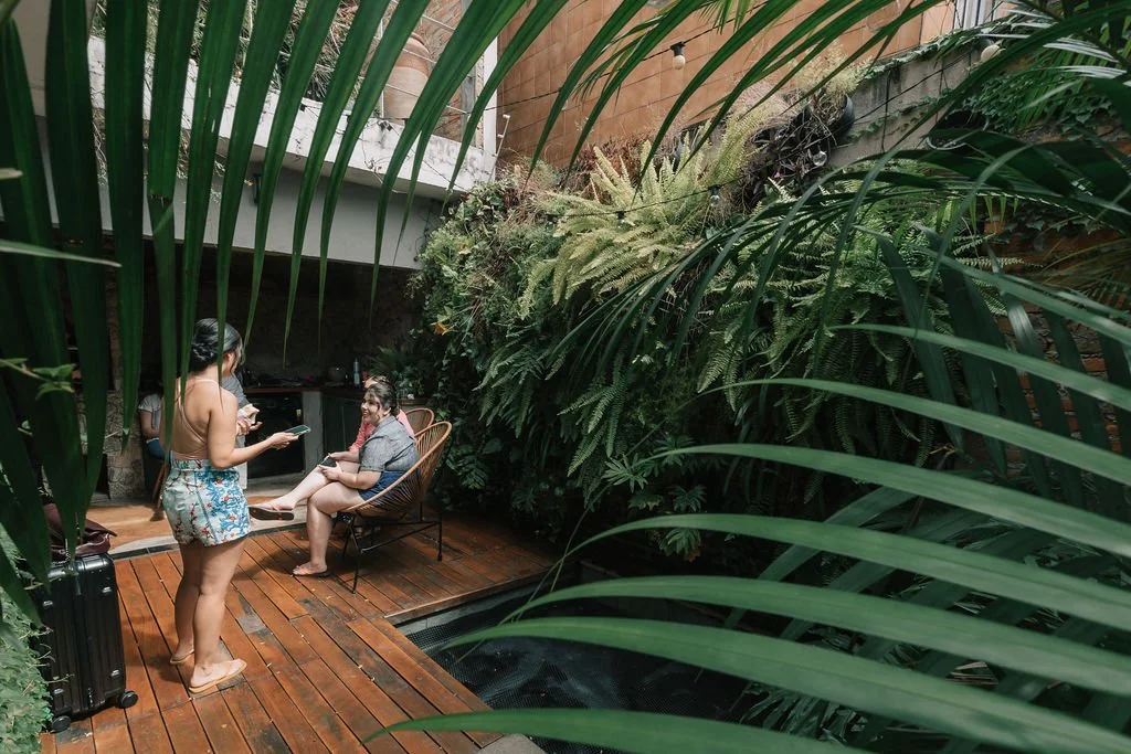 Duas mulheres em um espaço com plantas verdes grandes, uma sentada em uma cadeira com sobriedade e a outra de pé usando roupas de banho estampadas, ambas usando celulares.
