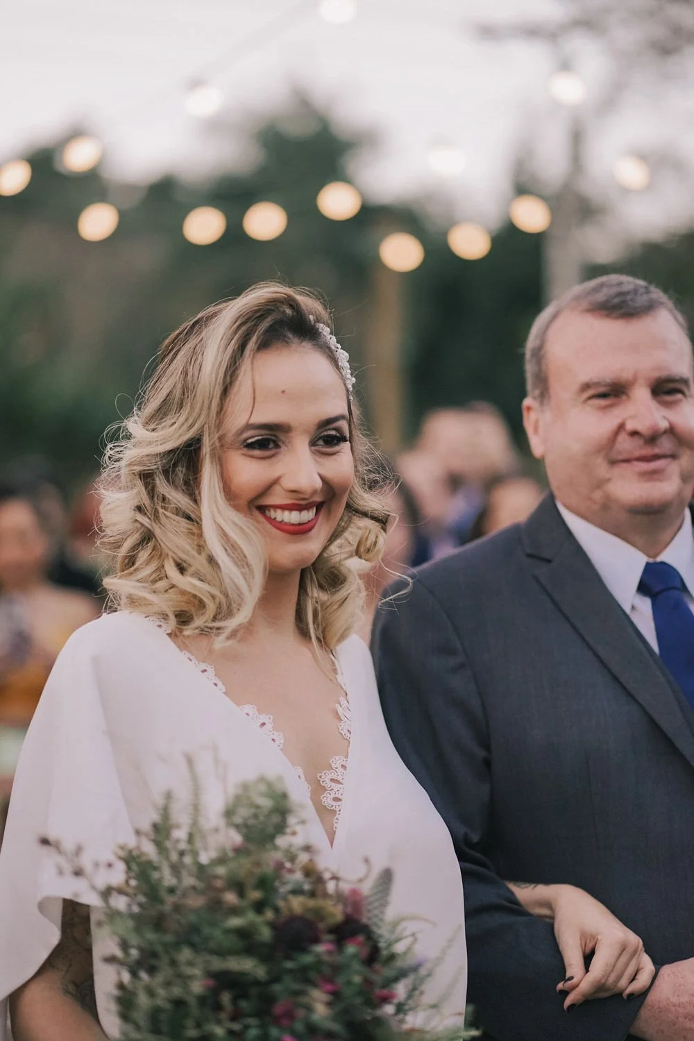 Mulher sorrindo usando vestido de casamento, ao lado de um homem em traje formal, em evento ao ar livre com luzes decorativas ao fundo.