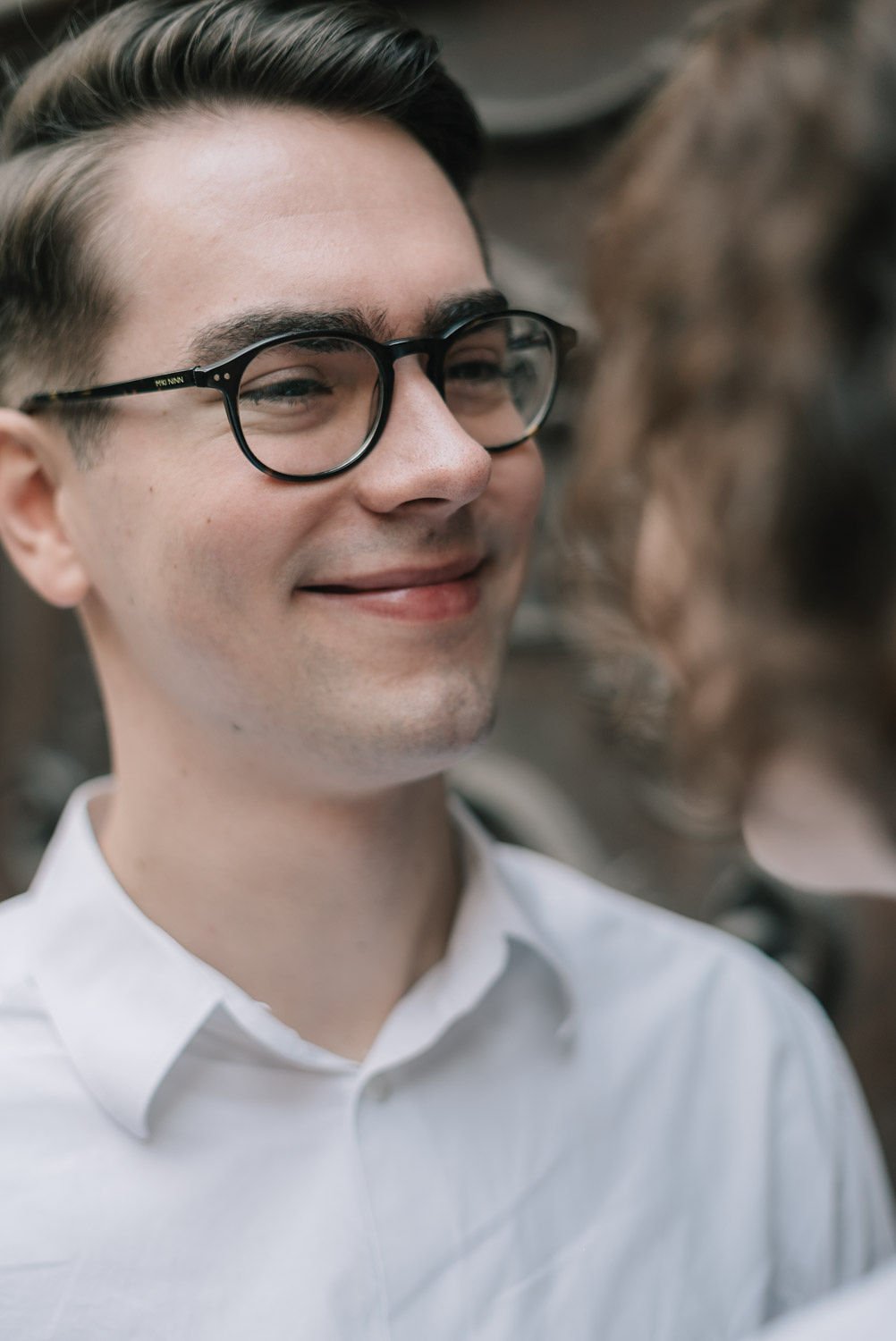 Homem sorridente usando óculos de armação preta e camisa branca, com uma mulher ao fundo.