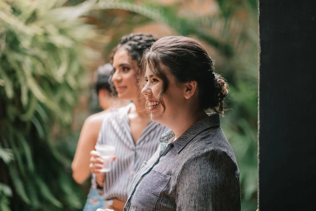 Duas mulheres sorrindo, uma com cabelo preso em um coque e a outra com cabelo cacheado, em um ambiente com plantas ao fundo.