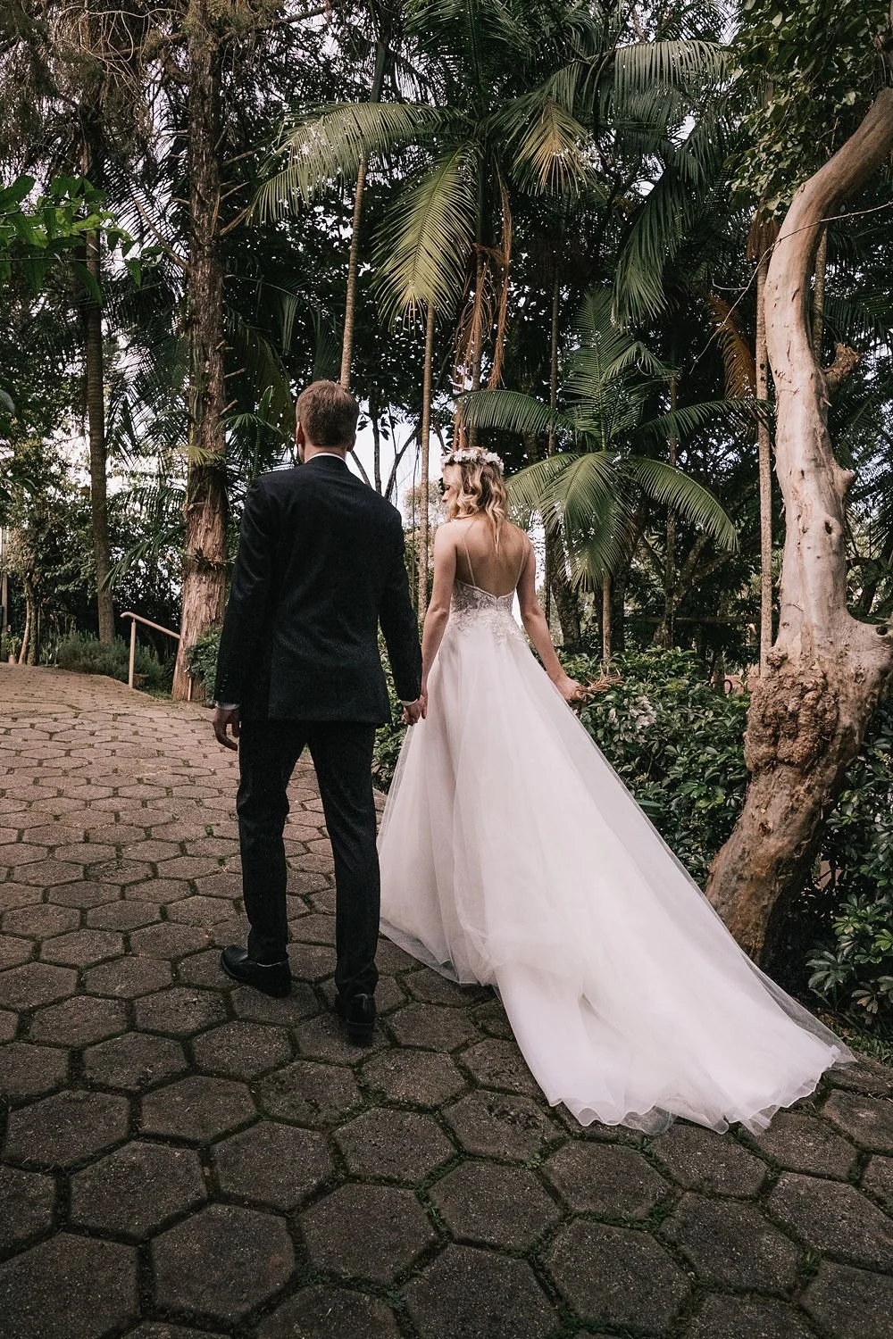 Casal de noivos caminhando de mãos dadas em um caminho de pedra, rodeados por árvores e plantas tropicais, durante um casamento ao ar livre.