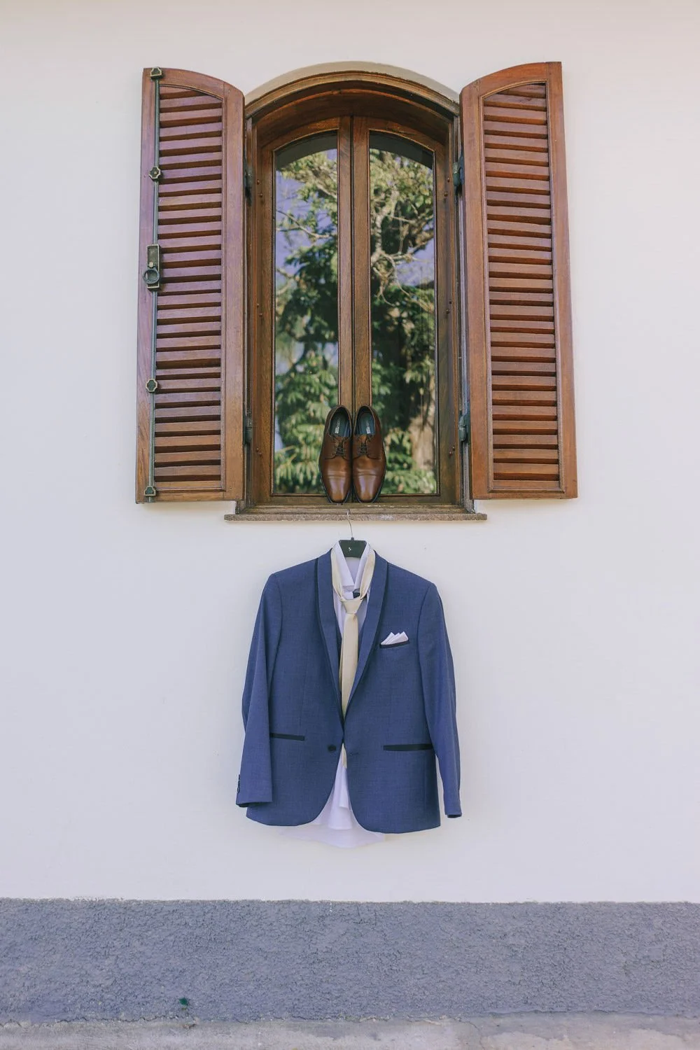 Casaco social azul, gravata branca, sapatos de couro marrom, tudo pendurado na parede baixa com janela de madeira aberta ao fundo, com vista para árvores verdes.
