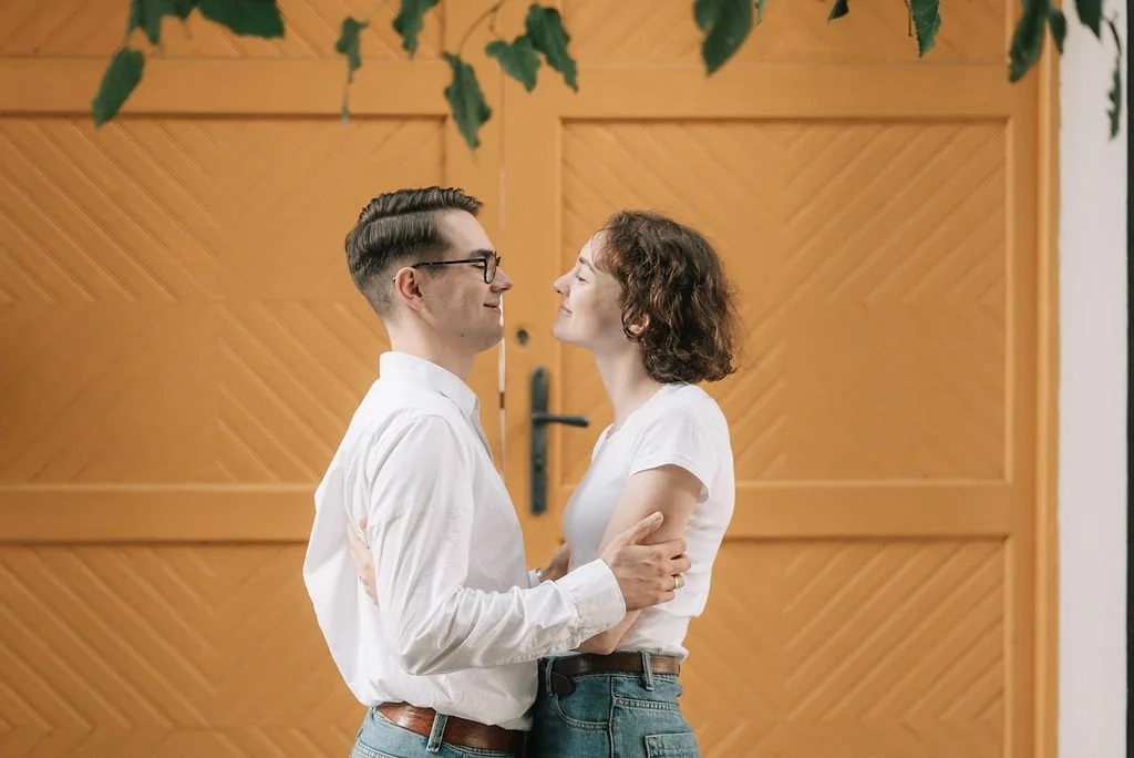 Um casal se olha fixamente, segurando-se um ao outro, em frente a uma porta de madeira com detalhes em madeira diagonal.