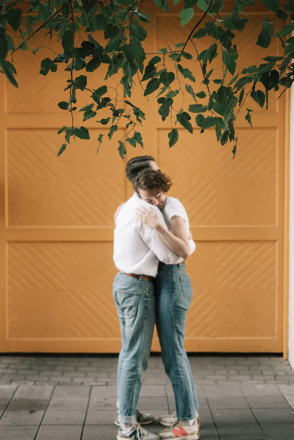 Um casal se abraçando carinhosamente em frente a uma parede de madeira, com folhas de planta penduradas acima.