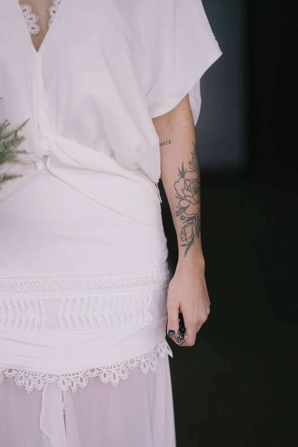 Braço de uma pessoa com tatuagem de flores e uma palavra, vestindo roupa branca com detalhes em renda.
