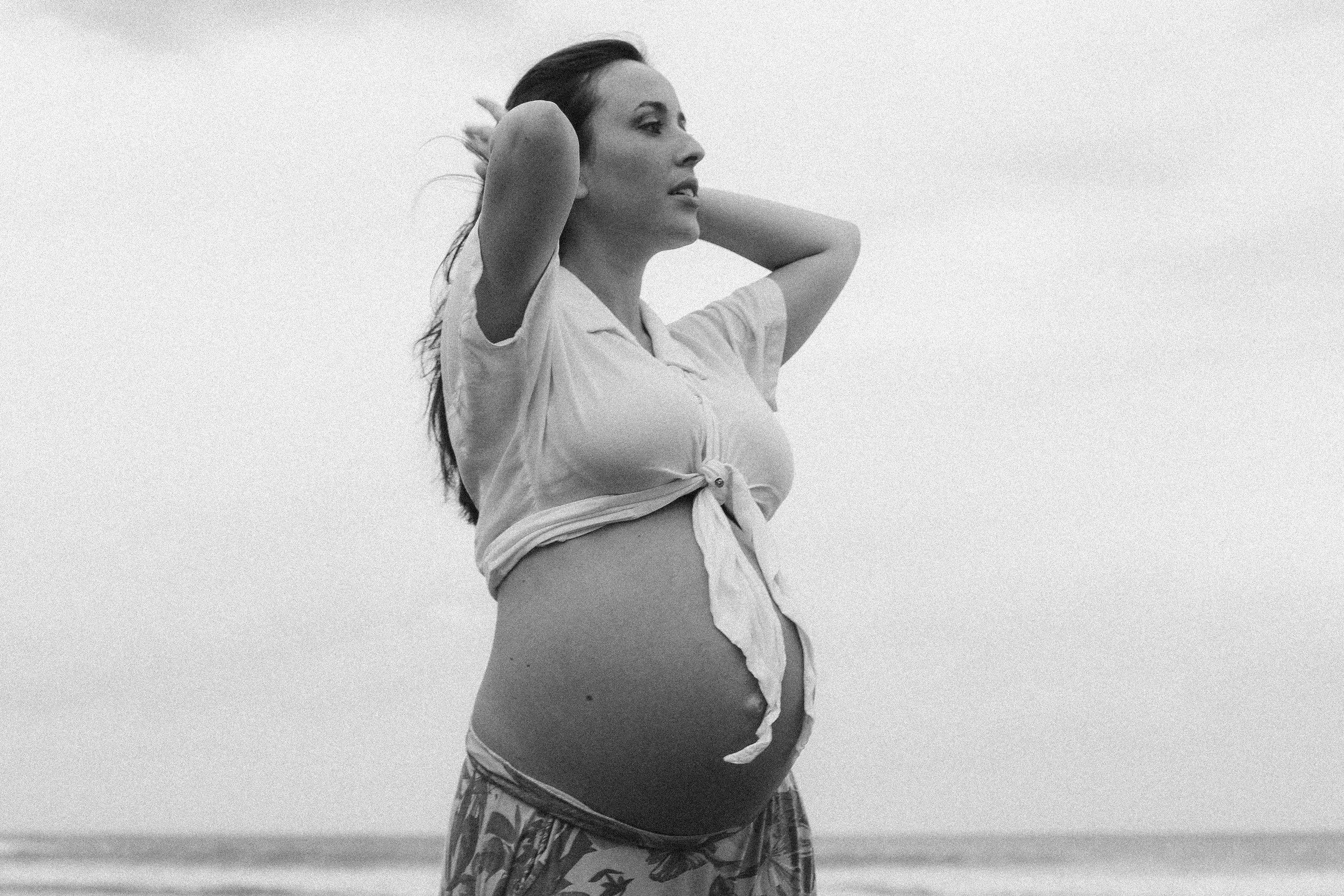 Mulher grávida de perfil na praia, com cabelo ao vento, usando camisa amarrada na frente e saia com estampa tropical.