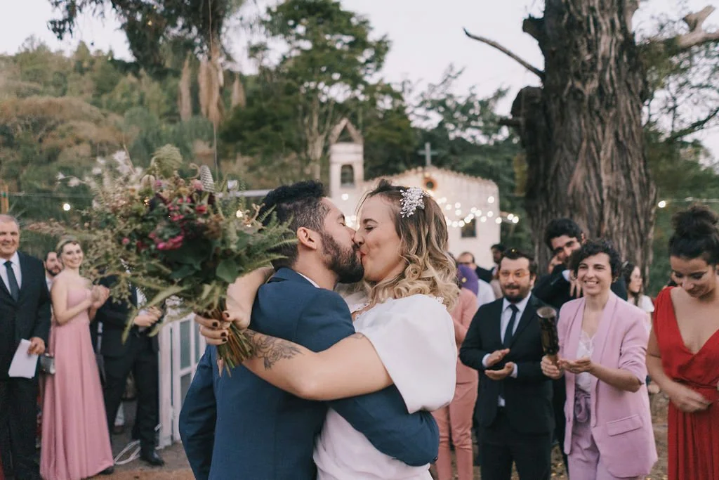 Casal se beijando em celebração de casamento ao ar livre com convidados sorrindo ao redor.