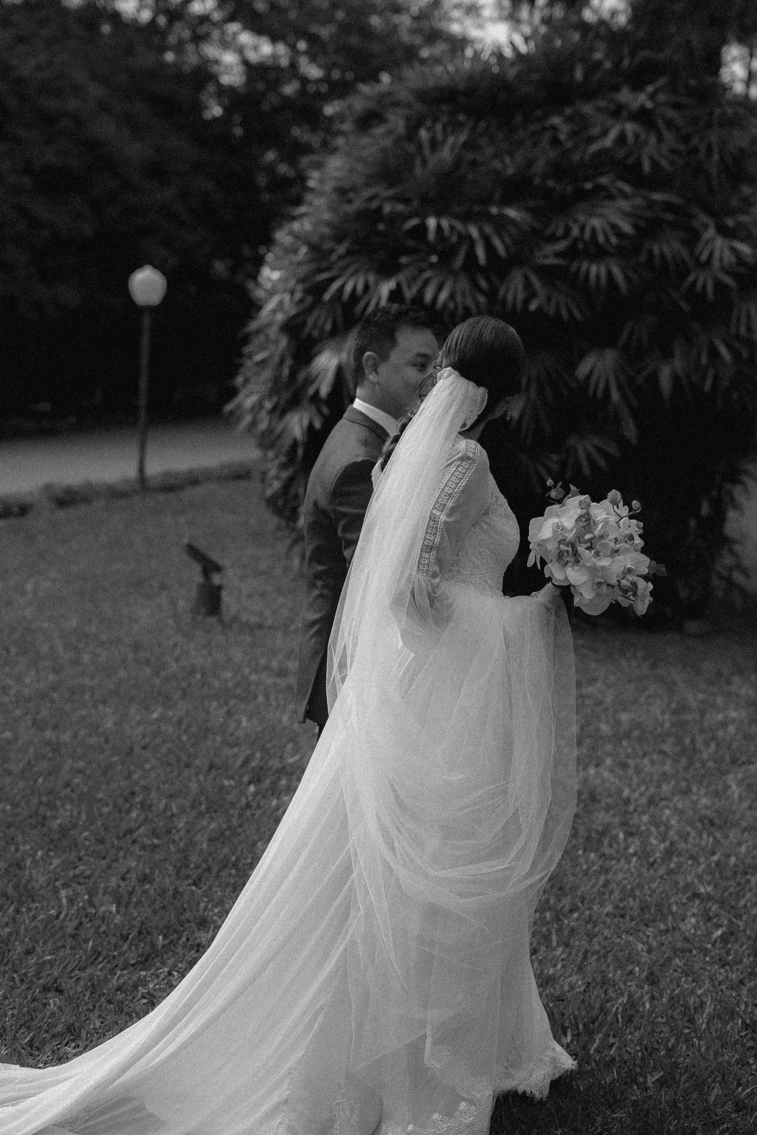 Casal de noivos de casamento ao ar livre, com a noiva segurando um buquê de flores e usando vestido longo, e o noivo usando terno, em tom preto e branco.