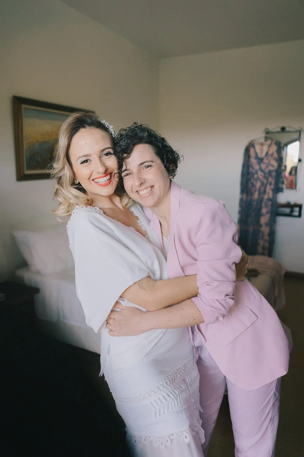 Duas mulheres sorrindo e se abraçando dentro de um quarto, uma vestindo um vestido branco e a outra com um casaco lilás.