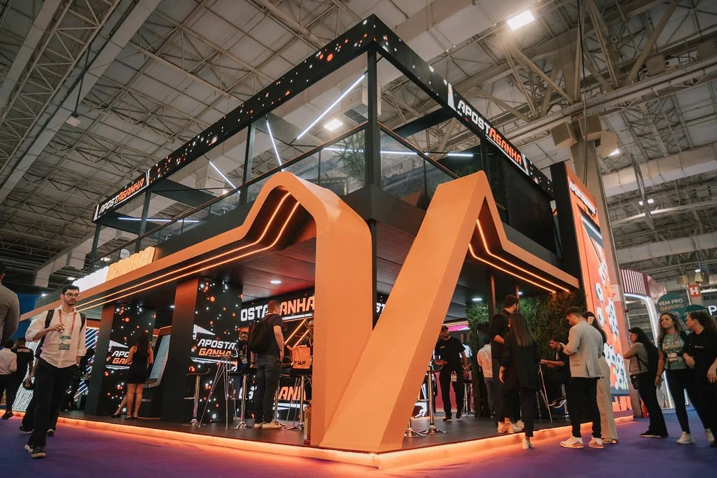 Estande de feira com estrutura moderna em cores preta e laranja, iluminação de LED, varias pessoas interagindo, ambiente de evento com teto alto e iluminação overhead.