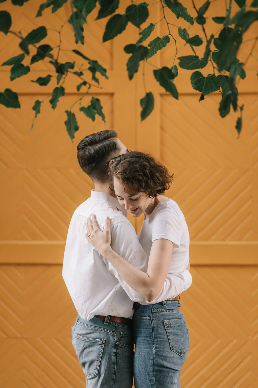 Um casal se abraçando dentro de uma sala com parede de madeira e folhas verdes penduradas no teto.