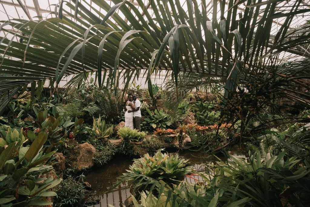 Casal andando na vegetação de um jardim ou estufa com plantas diversas ao redor.