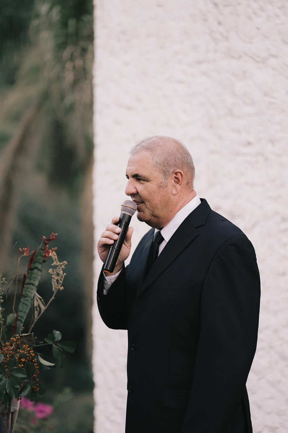 Homem idoso vestido com terno preto segurando microfone em um evento formal.