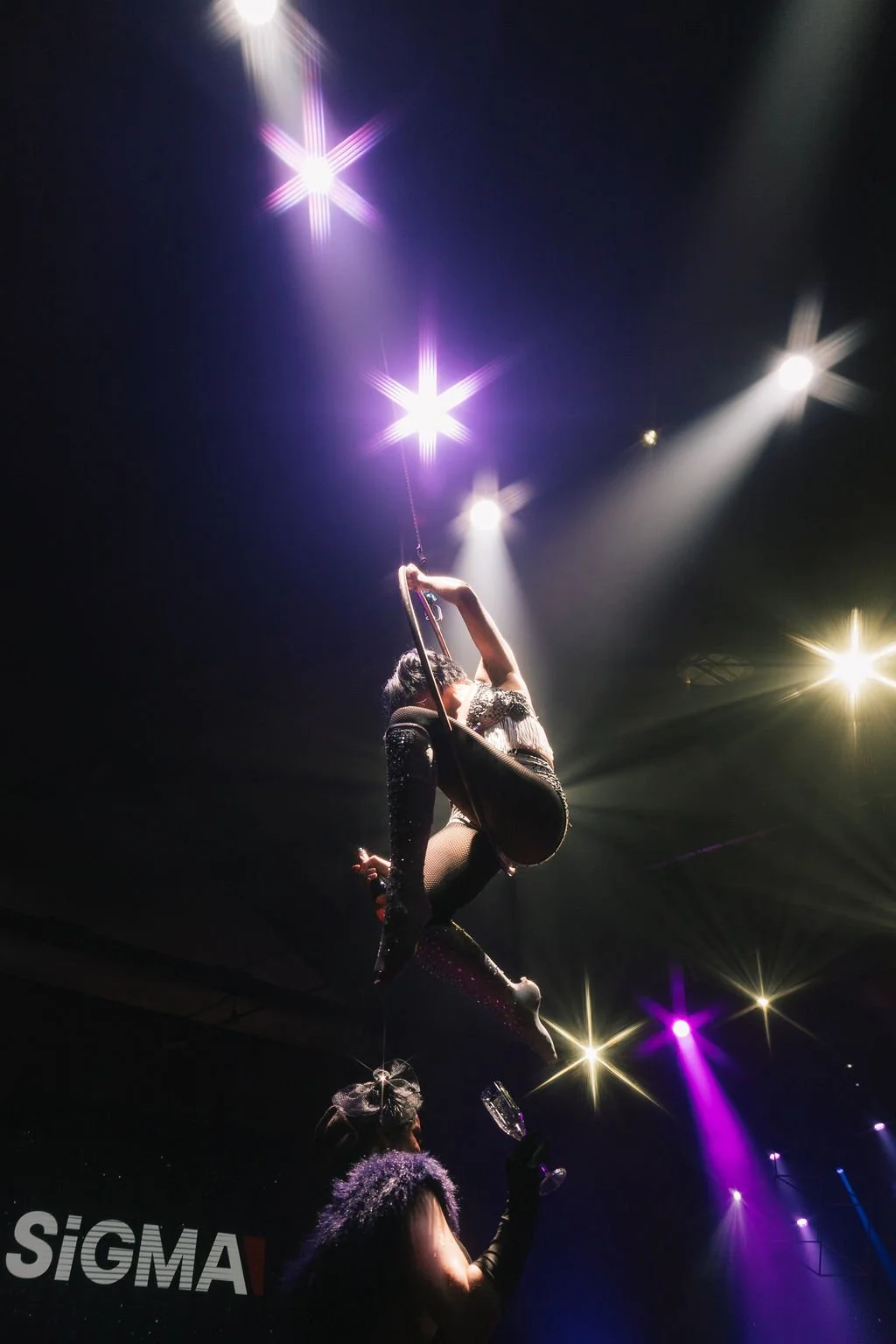 Um espetáculo de performance de arte em um palco com iluminação dramática, onde uma artista equilibra-se em uma hala, usando roupas brilhantes e acessórios, enquanto uma pessoa segura uma taça de vinho.