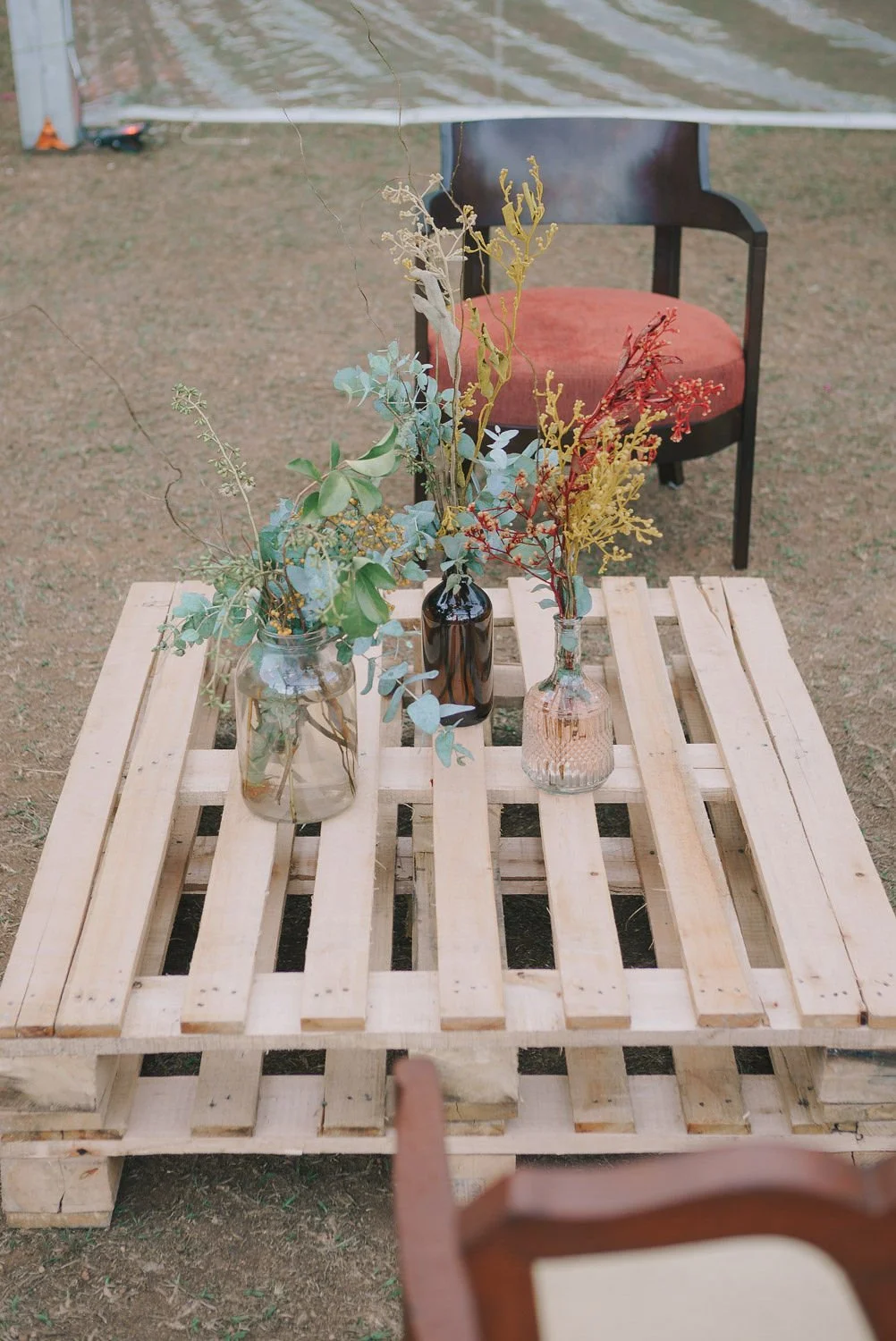 Mesa de paletes de madeira com três vasos de vidro com plantas e galhos decorativos, ao fundo uma cadeira preta com assento vermelho